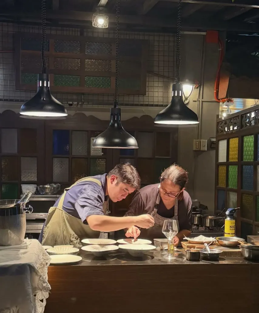Two chefs plating side by side at a kitchen counter under hanging lamps and capiz-style colored glass